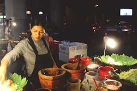 A street vendor is preparing food at an outdoor market stall. She is focused and wearing a gray sweater with a black apron. Various ingredients are spread out on the counter, including leafy greens, mortar and pestle, and several bowls. The setting is dimly lit, with a small lamp providing illumination.