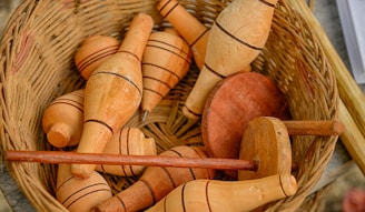 A wicker basket contains several wooden spinning tops, each with a pointed end and decorated with black lines. The objects display a smooth finish and are arranged in a somewhat haphazard manner within the basket.