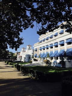 Buildings with white exteriors and blue awnings are aligned along a tidy, green lawn. A sidewalk runs parallel to the buildings, with trees providing shade. The sky is clear and blue. The main building is marked with 'Buckstaff Baths.'