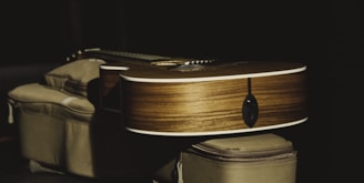 An acoustic guitar lies horizontally across a soft, cushion-like surface. The guitar's wooden body contrasts with the dark background, with light reflecting off its polished surface.