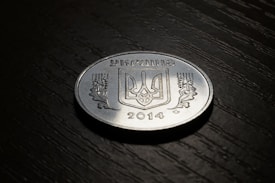 A coin placed on a dark textured surface. The coin features the coat of arms of Ukraine in the center with the word 'Україна' inscribed above it and '2014' written below. There are also decorative elements on both sides of the coat of arms.