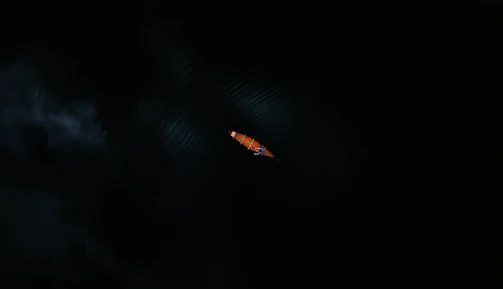 A single, small kayak is seen from above, cutting through dark waters. The kayak is orange, and the water around it is nearly black, creating a stark contrast. Minimal light reflects off the surface, emphasizing the solitude and tranquility of the scene.