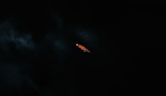 A single, small kayak is seen from above, cutting through dark waters. The kayak is orange, and the water around it is nearly black, creating a stark contrast. Minimal light reflects off the surface, emphasizing the solitude and tranquility of the scene.