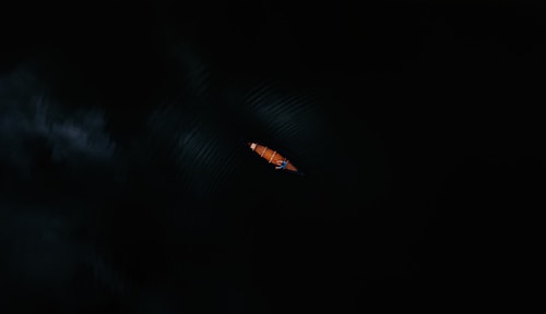 A single, small kayak is seen from above, cutting through dark waters. The kayak is orange, and the water around it is nearly black, creating a stark contrast. Minimal light reflects off the surface, emphasizing the solitude and tranquility of the scene.
