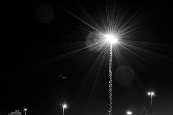 A brightly illuminated streetlight stands tall against a dark night sky. Its light radiates in starburst patterns, casting flares and reflections around it. Smaller streetlights in the background contribute to a soft, dispersed glow.