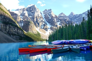 Colorful canoes are lined up on a pristine blue lake, with snow-capped mountains rising majestically in the background. Dense, green forests border the lake, creating a serene and natural setting.