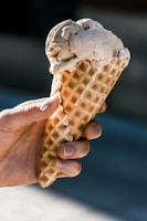 A hand is holding a waffle cone with a scoop of creamy ice cream on top. The ice cream has a light brown hue, and its texture appears smooth and slightly melting. The person's fingers are visible, gripping the cone, and the background is softly blurred.