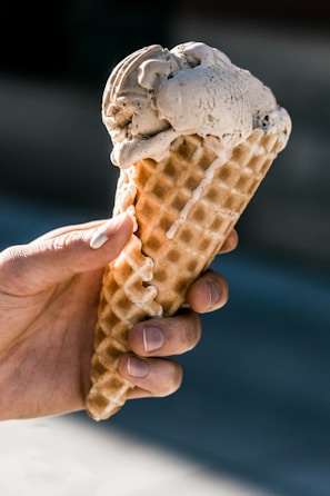 A hand is holding a waffle cone with a scoop of creamy ice cream on top. The ice cream has a light brown hue, and its texture appears smooth and slightly melting. The person's fingers are visible, gripping the cone, and the background is softly blurred.
