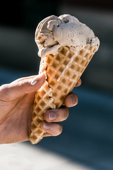 A hand is holding a waffle cone with a scoop of creamy ice cream on top. The ice cream has a light brown hue, and its texture appears smooth and slightly melting. The person's fingers are visible, gripping the cone, and the background is softly blurred.