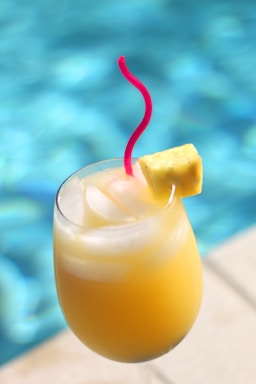 A refreshing tropical drink served in a clear glass with ice cubes and a slice of pineapple on the rim, complemented by a bright red straw. The background features a swimming pool with shimmering blue water.