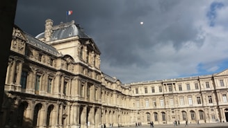 A grand historical building with intricate architectural details and a French flag on top under a cloudy sky. There are a few people walking in the open courtyard.