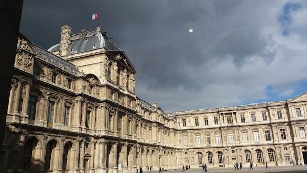 A grand historical building with intricate architectural details and a French flag on top under a cloudy sky. There are a few people walking in the open courtyard.