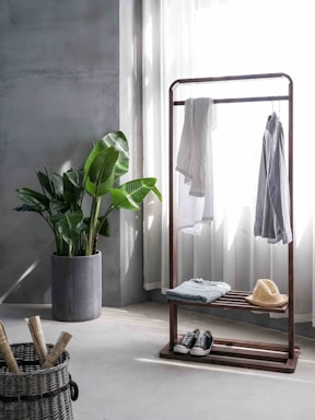 A minimalist room with a clothing rack holding a light shirt and a white garment. Below, there's a folded item and a beige straw hat. A pair of sneakers is placed on the floor. A large potted plant with broad green leaves sits beside the rack. A woven basket contains rolled items, and light filters through sheer curtains, casting soft shadows on the neutral-toned walls.