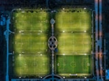 An aerial view of multiple illuminated soccer fields at night, arranged in a grid layout with visible field markings and goal posts. Surrounding the fields are pathways and a small circular area in the center. There are small groups of people playing on several fields. Adjacent parking areas and streets are faintly visible on the sides.