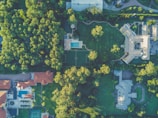 An aerial view of a residential area featuring large houses surrounded by lush green trees. The image shows a swimming pool, a soccer field, and various rooftops with red and gray tiles. Winding pathways and driveways intermingle within the landscaped gardens.