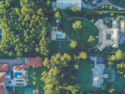 An aerial view of a residential area featuring large houses surrounded by lush green trees. The image shows a swimming pool, a soccer field, and various rooftops with red and gray tiles. Winding pathways and driveways intermingle within the landscaped gardens.