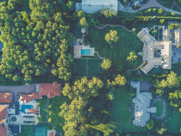 An aerial view of a residential area featuring large houses surrounded by lush green trees. The image shows a swimming pool, a soccer field, and various rooftops with red and gray tiles. Winding pathways and driveways intermingle within the landscaped gardens.