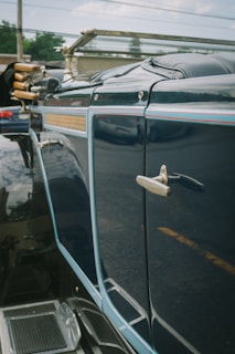 A close-up view of a vintage car with a glossy blue finish and intricate detailing, including a metal handle and trim. The reflection of the surroundings is visible on the car's surface, adding to the antique aesthetic.