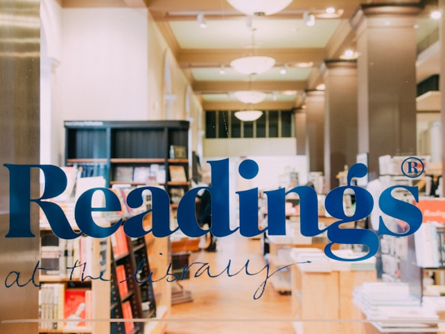 A bookstore interior is seen through a glass door with blue lettering spelling 'Readings'. Shelves filled with books and display stands are visible. The lighting is warm and ambient, creating a welcoming atmosphere.