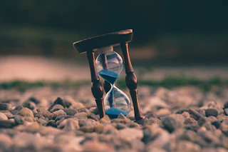 Wooden hourglass with blue sand mid-fall, tilted among smooth grey and tan pebbles
