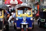 A colorful tuk-tuk parked in a bustling street setting. The tuk-tuk is blue with yellow trim and displays a 'TAXI' sign on its roof. A Coca-Cola umbrella and various hanging items are in the background, suggesting a vibrant street market environment.