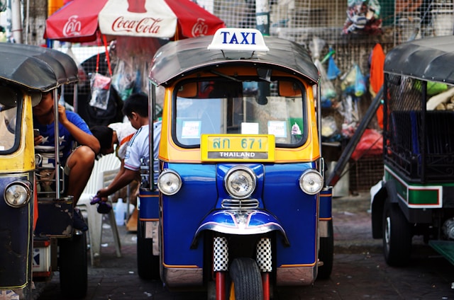 A colorful tuk-tuk parked in a bustling street setting. The tuk-tuk is blue with yellow trim and displays a 'TAXI' sign on its roof. A Coca-Cola umbrella and various hanging items are in the background, suggesting a vibrant street market environment.
