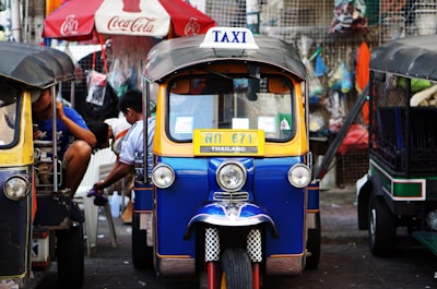 A colorful tuk-tuk parked in a bustling street setting. The tuk-tuk is blue with yellow trim and displays a 'TAXI' sign on its roof. A Coca-Cola umbrella and various hanging items are in the background, suggesting a vibrant street market environment.