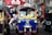 A colorful tuk-tuk parked in a bustling street setting. The tuk-tuk is blue with yellow trim and displays a 'TAXI' sign on its roof. A Coca-Cola umbrella and various hanging items are in the background, suggesting a vibrant street market environment.