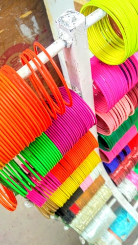 A variety of colorful bangles are neatly arranged on a white rack. The bangles range in color from vibrant neon yellow to green, pink, red, and orange. Each set is tightly grouped together creating a visually striking and organized display.