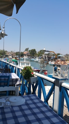 A peaceful outdoor dining area with blue and white checkered tablecloths is situated next to a body of water. The area features decorative plants and nautical elements like buoys and ropes. In the background, there are seated guests, small boats moored, and a crane arm visible, suggesting a harbor setting. The sky is clear, and the overall scene conveys a relaxed seaside atmosphere.