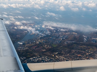 The image captures an aerial view from an airplane over a coastal city, showing a mix of urban and industrial landscapes with sprawling buildings, factories, and a water body likely connected to the sea. Clouds are scattered throughout the sky, and the airplane wing is visible on the left side of the image.