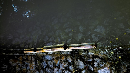 Two reflective pipes extend horizontally across a surface, possibly part of a water or drainage system. The background consists of natural stones arranged in a wall, with some vegetation growing around it. The lighting creates a contrast between the dark and light areas, highlighting the textures of the stones.
