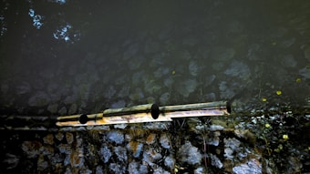 Two reflective pipes extend horizontally across a surface, possibly part of a water or drainage system. The background consists of natural stones arranged in a wall, with some vegetation growing around it. The lighting creates a contrast between the dark and light areas, highlighting the textures of the stones.