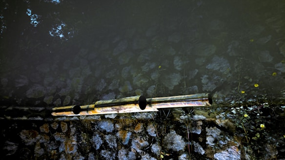 Two reflective pipes extend horizontally across a surface, possibly part of a water or drainage system. The background consists of natural stones arranged in a wall, with some vegetation growing around it. The lighting creates a contrast between the dark and light areas, highlighting the textures of the stones.
