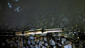 Two reflective pipes extend horizontally across a surface, possibly part of a water or drainage system. The background consists of natural stones arranged in a wall, with some vegetation growing around it. The lighting creates a contrast between the dark and light areas, highlighting the textures of the stones.