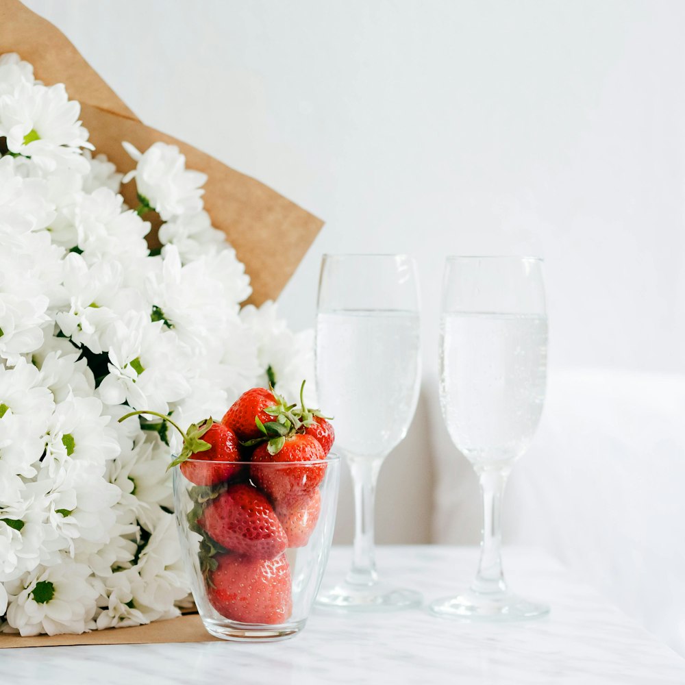 tasse en verre remplie de fraises rouges à côté du bouquet de fleurs blanches