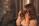 A person with long hair in a patterned shirt is holding a Nikon camera up to their face, suggesting they are about to take a photograph. The background includes a backpack and a metallic, textured surface, with warm lighting creating a cozy atmosphere.
