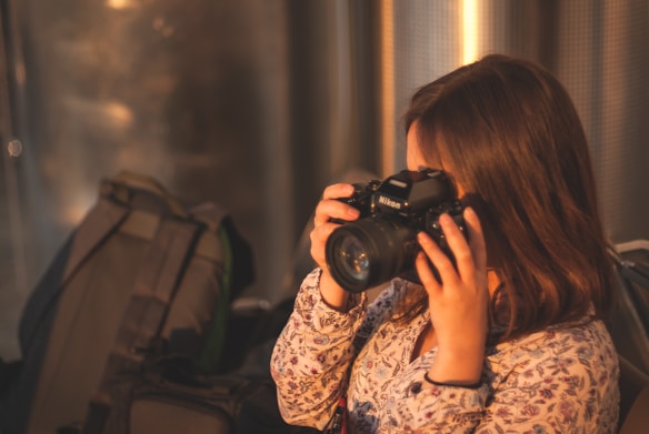 A person with long hair in a patterned shirt is holding a Nikon camera up to their face, suggesting they are about to take a photograph. The background includes a backpack and a metallic, textured surface, with warm lighting creating a cozy atmosphere.