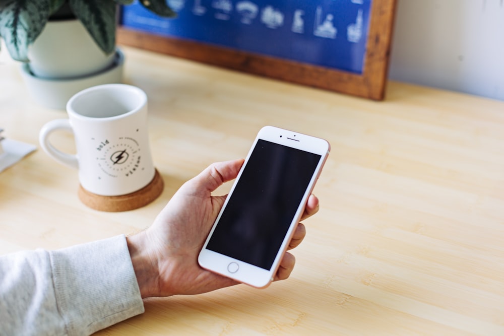 A person holding an iPhone over a desk with a white mug