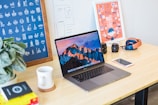 A neatly arranged workspace features a laptop displaying a mountain landscape, positioned centrally on a wooden desk. Surrounding the laptop are various items including a potted plant, a coffee mug on a coaster, a smartphone, camera lenses, a pair of headphones, and design posters on the wall. The environment conveys a creative and organized atmosphere.