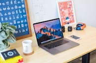 A neatly arranged workspace features a laptop displaying a mountain landscape, positioned centrally on a wooden desk. Surrounding the laptop are various items including a potted plant, a coffee mug on a coaster, a smartphone, camera lenses, a pair of headphones, and design posters on the wall. The environment conveys a creative and organized atmosphere.