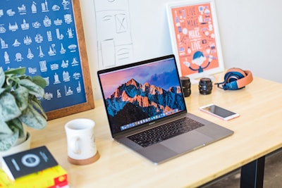 A neatly arranged workspace features a laptop displaying a mountain landscape, positioned centrally on a wooden desk. Surrounding the laptop are various items including a potted plant, a coffee mug on a coaster, a smartphone, camera lenses, a pair of headphones, and design posters on the wall. The environment conveys a creative and organized atmosphere.