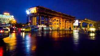 A construction site at night with a large, illuminated structure surrounded by scaffolding. Vehicles are seen passing by on a wet road reflecting the lights. In the background, there's a hospital building with brightly lit signage.