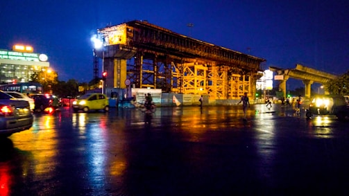 A construction site at night with a large, illuminated structure surrounded by scaffolding. Vehicles are seen passing by on a wet road reflecting the lights. In the background, there's a hospital building with brightly lit signage.