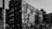 A black and white urban scene featuring a large mural on the side of a building, depicting a detailed and artistic portrayal of a person. Surrounding buildings have multiple stories with visible fire escapes, and several parked cars are seen below.