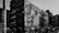 A black and white urban scene featuring a large mural on the side of a building, depicting a detailed and artistic portrayal of a person. Surrounding buildings have multiple stories with visible fire escapes, and several parked cars are seen below.