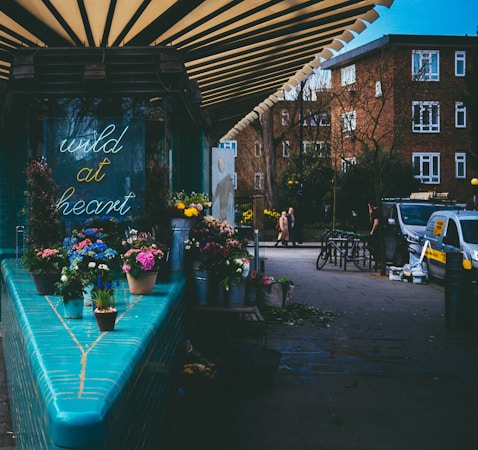 A small street floral shop features an inviting display of colorful flowers in pots and metal containers. A neon sign with the words 'wild at heart' is visible on the wall. The shop is under a striped awning. In the background, brick apartment buildings and parked vehicles are seen, while a few pedestrians walk along the sidewalk.