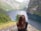 A person with long brown hair stands on a rocky cliff, overlooking a scenic fjord surrounded by steep, lush green mountains. The water is calm and a dark blue, stretching between the mountains. The landscape is tranquil and expansive.