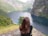 A person with long brown hair stands on a rocky cliff, overlooking a scenic fjord surrounded by steep, lush green mountains. The water is calm and a dark blue, stretching between the mountains. The landscape is tranquil and expansive.