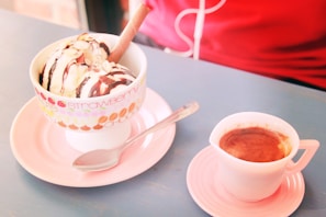 Two scoops of vanilla ice cream topped with chocolate syrup and almond slices are served in a colorful cup labeled with words like 'strawberry' and 'choco'. A wafer stick is placed in the ice cream. Next to it, there is a shot of espresso in a small pink cup on a matching saucer. A spoon lies beside the cup of ice cream on a pastel pink plate. The background features a red garment and a blue tabletop.